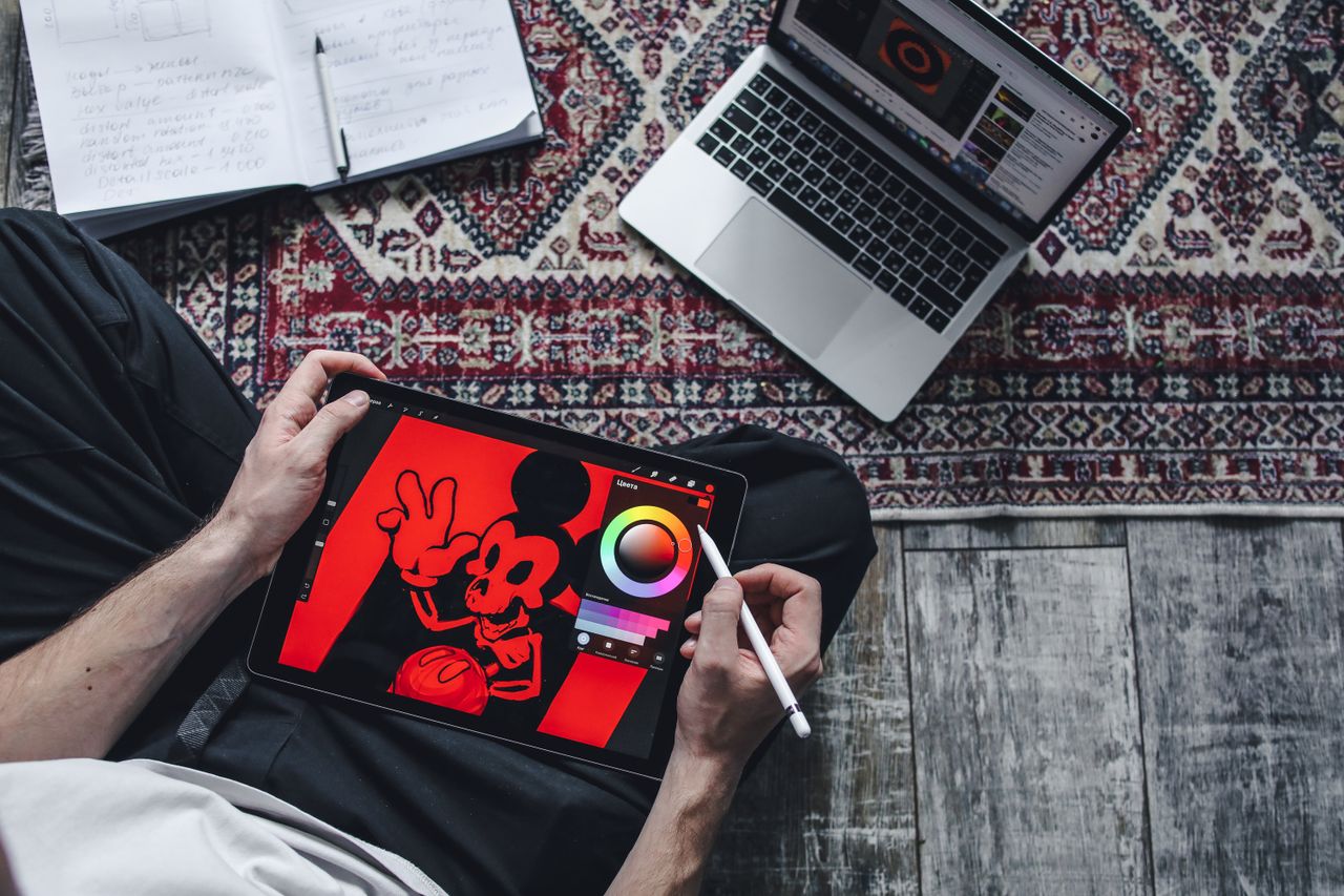 A person drawing on a digital tablet while seated on a patterned rug indoors.